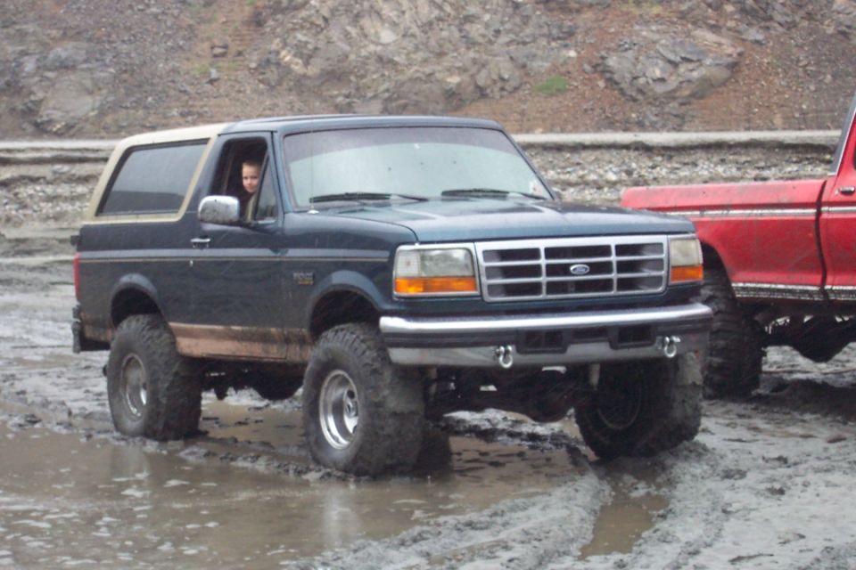Dark blue 1995 Ford Bronco with a tan top, lifted suspension, and large tires driving through mud. A red pickup truck is partially visible to the right