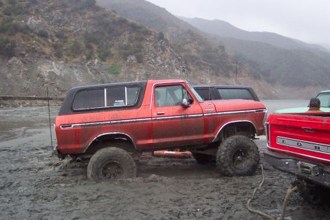 1995 Ford Bronco - A lifted red SUV is stuck in thick mud near a body of water with mountains in the background.