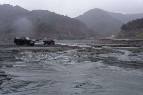 1995 Ford Bronco - Two SUVs drive through a desolate, muddy canyon under overcast skies.