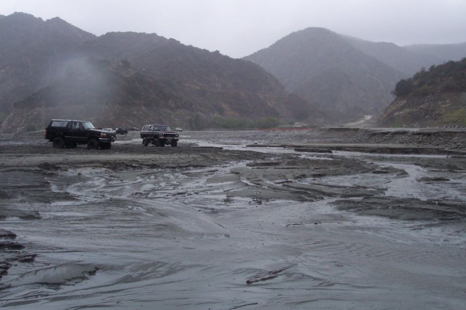 Two dark SUVs navigate a muddy, rocky riverbed with mountains in the background