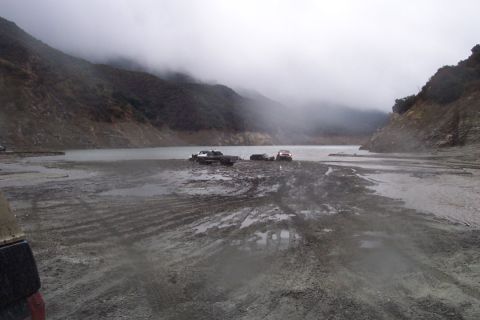 1995 Ford Bronco - Multiple vehicles are mired in mud near a lake, with mountains shrouded in fog in the background.