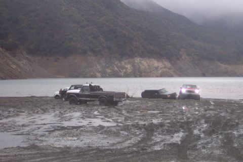 1995 Ford Bronco - Multiple vehicles are stuck in the mud by a lake on a cloudy day.