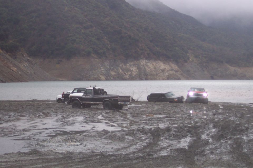 Several vehicles stuck in mud near a body of water with a hill in the background