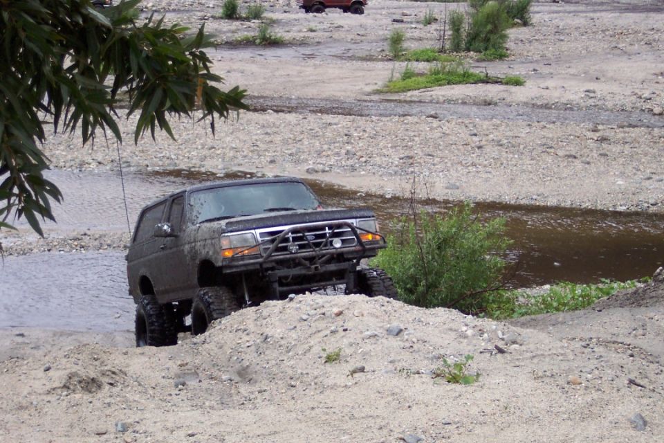 1995 Ford Bronco climbing a dirt mound in a rocky stream bed. Another vehicle is visible in the background