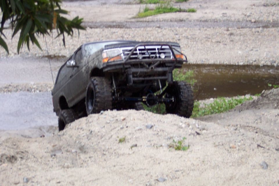 Off-road SUV with large tires climbing a dirt hill next to water. Custom front bumper and roll cage visible