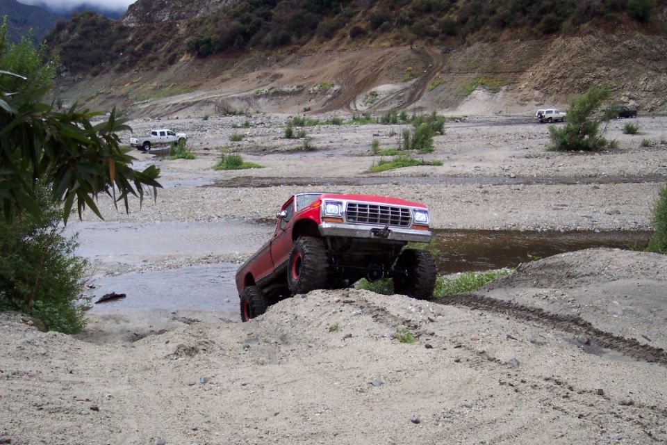 Red lifted pickup truck with large tires driving through a rocky stream bed. Other vehicles are visible in the background