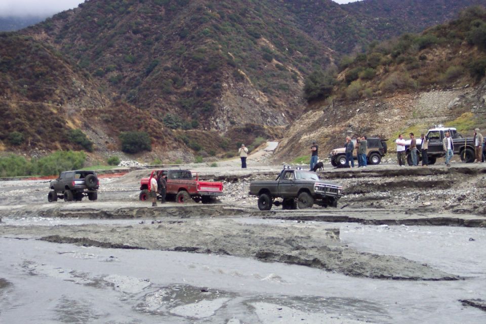 Several off-road trucks and SUVs navigate a muddy, rocky terrain with steep hills in the background