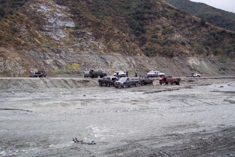 1995 Ford Bronco - Several off-road vehicles are gathered in a dry riverbed near a steep, rocky mountainside.