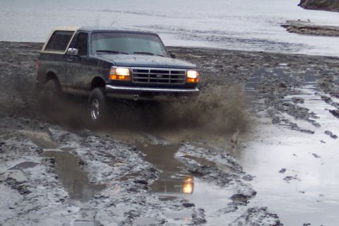 1995 Ford Bronco - A Ford Bronco navigates a challenging muddy terrain, kicking up spray.