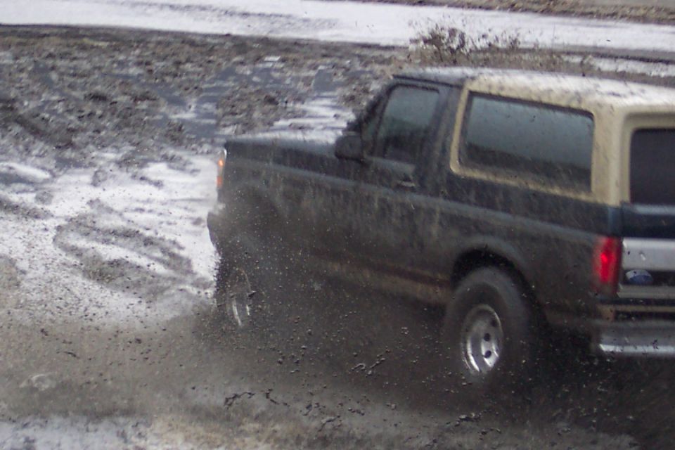 Dark Ford Bronco with tan top driving through mud, rear quarter view