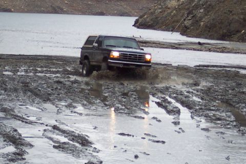 1995 Ford Bronco - A dark green 1995 Ford Bronco navigates a muddy terrain near a body of water.