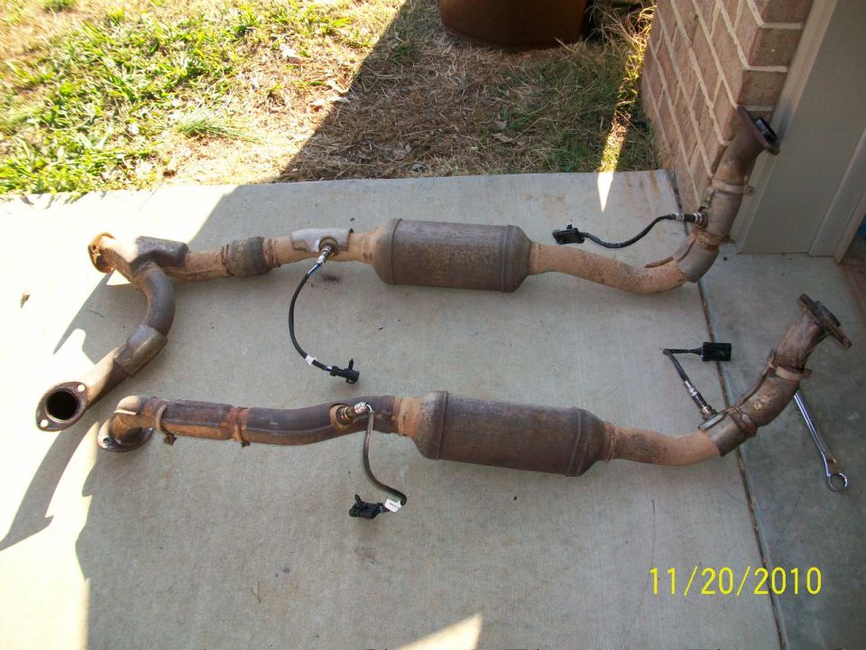 Two rusty exhaust systems with catalytic converters and oxygen sensors laid out on concrete