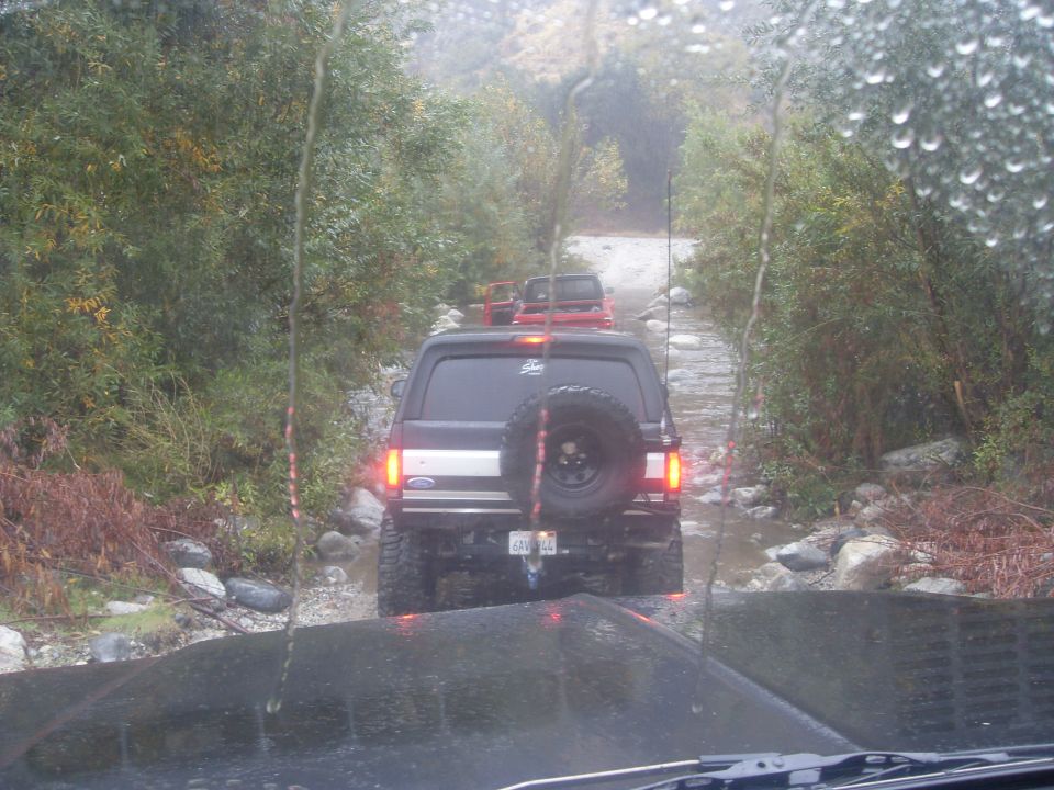 Several SUVs driving on a rocky, wet trail. The lead vehicle is a black Ford Bronco with a spare tire on the back