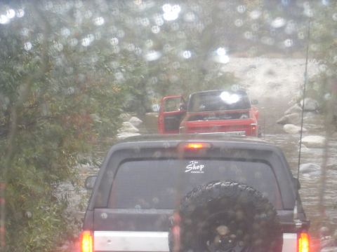 1979 Ford Bronco - Two vehicles navigate a shallow, rocky waterway surrounded by trees.