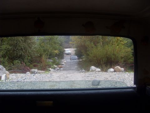 1979 Ford Bronco - Two SUVs navigate a rough dirt road surrounded by trees and rocks, seen from the perspective of another vehicle.