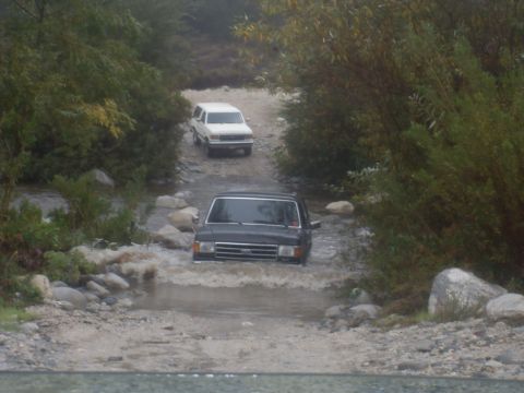 1979 Ford Bronco - Two SUVs navigate a rocky stream bed surrounded by trees.