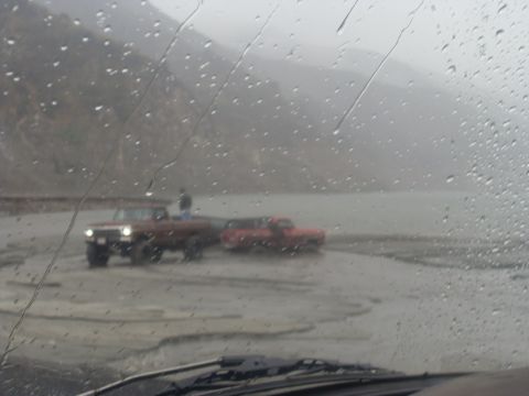 1979 Ford Bronco - Two trucks navigate a muddy terrain under rainy conditions, with a mountainous backdrop.