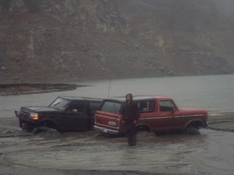 1979 Ford Bronco - Two SUVs, a red 1979 Ford Bronco and a black model, are stuck in the mud near a body of water.