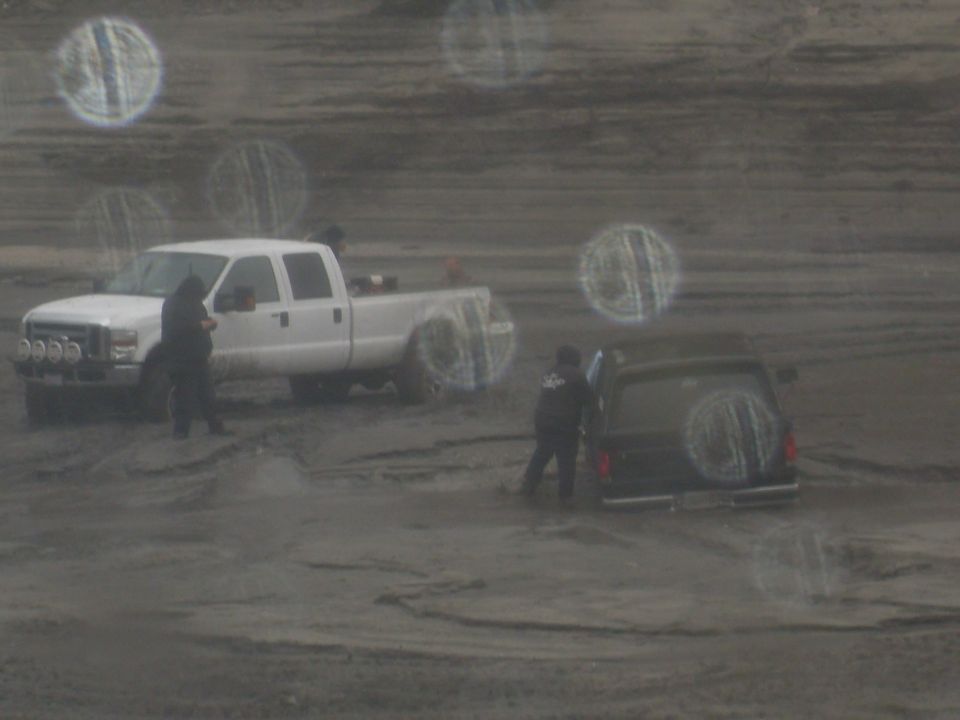 White pickup truck and black SUV parked in a muddy, off-road area with two people standing nearby