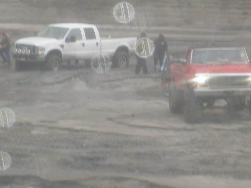Two pickup trucks, one white crew cab and one red, are in a muddy area with people around them