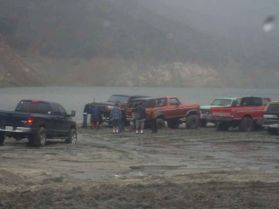 Group of trucks and SUVs gathered on a muddy shore near water, with a 1979 Ford Bronco among them