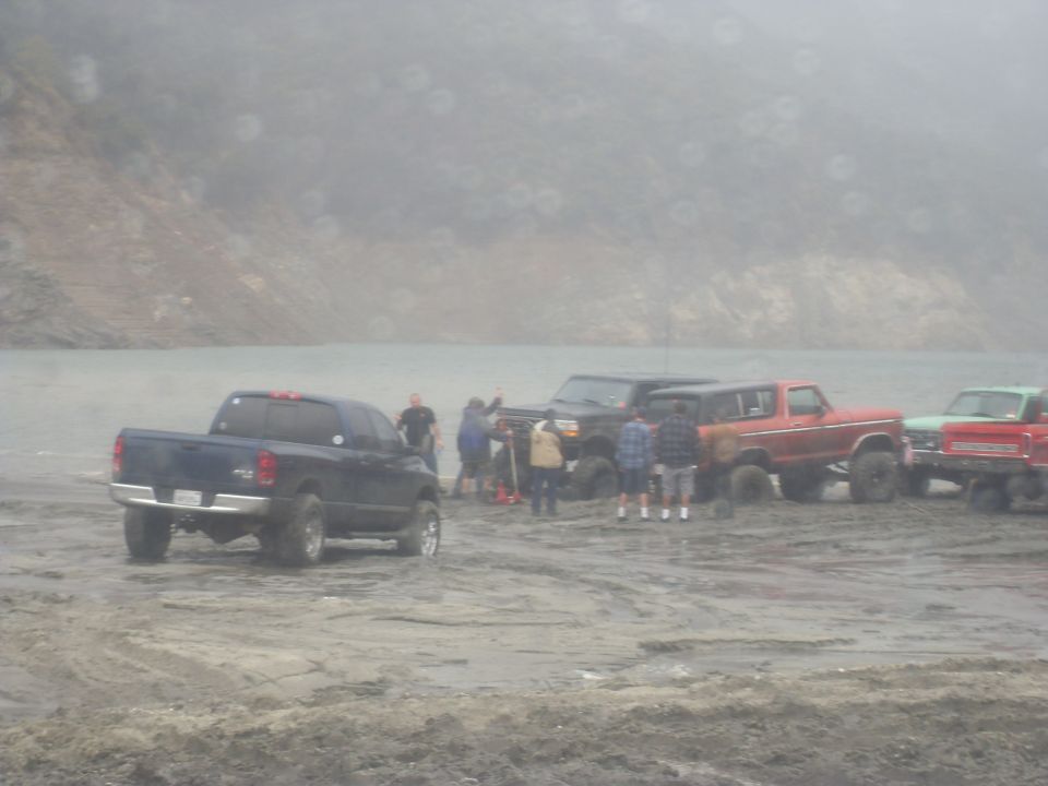 Several trucks and SUVs gathered on a sandy area near water, with people standing around them