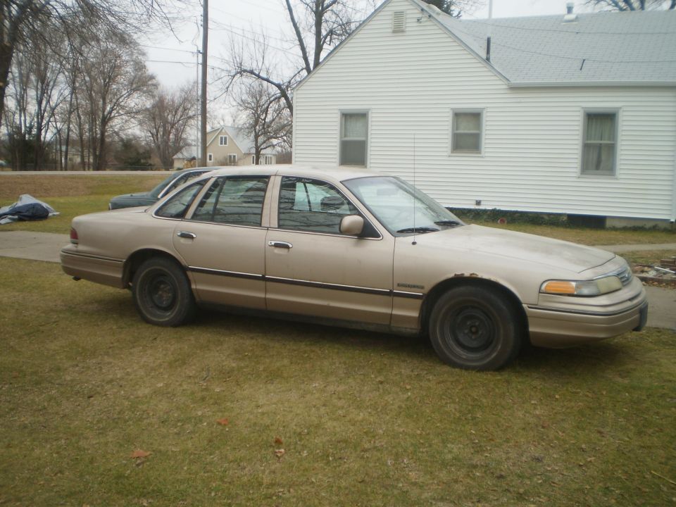 Tan Ford Crown Victoria sedan with steel wheels parked on grass by a white house