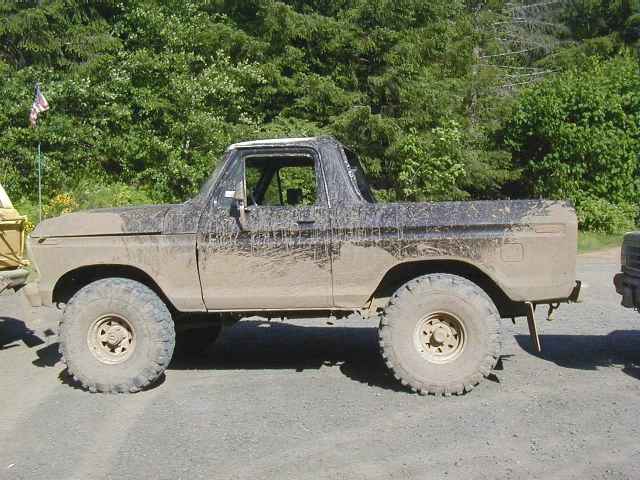 Mud-covered lifted Ford Bronco, top removed, on large tires parked on gravel.