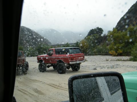 1982 Ford Bronco - A red 1982 Ford Bronco is seen from behind, driving on a dirt road with other vehicles in a mountainous, rainy landscape.