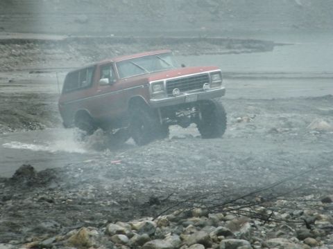 1982 Ford Bronco - This red SUV is navigating a rough, wet terrain with large tires kicking up water.