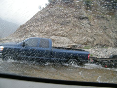 1982 Ford Bronco - Pickup truck and trailer navigate flooded road conditions near a rocky slope.