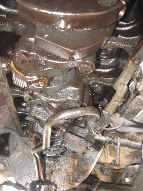 1981 Ford Bronco - Oily under-hood shot looking down the front of an engine block at the mechanical fuel pump and its steel fuel line, with the oil pan rail a…