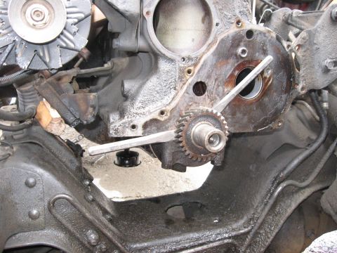 1981 Ford Bronco - Front of an engine with the timing cover off and camshaft gear removed, showing the crankshaft snout gear held with a long wrench and the e…