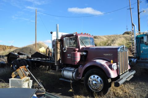 1959 Kenworth W900A - A maroon Kenworth semi-truck cab and chassis sits in a yard next to another truck under a blue sky.
