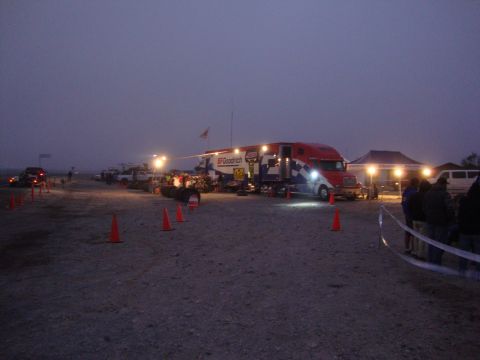 1990 Ford Bronco - BFGoodrich semi-truck support rig and lit tent at a dusty desert race staging area at dusk, marked by traffic cones.