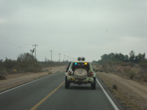 1990 Ford Bronco - camouflage-painted SUV with a rear-mounted spare tire drives down a paved desert highway lined with cacti and power poles.