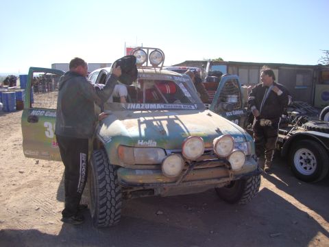 1990 Ford Bronco - crew tends to a mud-caked green off-road race truck marked 303 with Hella lights in a dusty desert pit area.