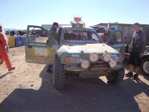 1990 Ford Bronco - weathered green off-road race truck numbered 303, covered in sponsor and SCORE decals, sits with its door open as men stand nearby.