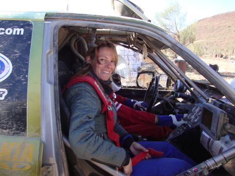 1990 Ford Bronco - smiling woman in a red racing suit sits behind the wheel inside the gutted cockpit of a desert race vehicle.