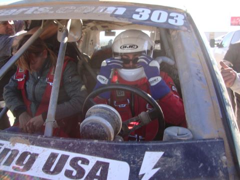 1990 Ford Bronco - helmeted driver in a red racing suit grips the steering wheel inside a race vehicle marked 303, with a passenger beside.