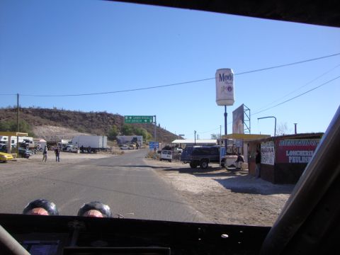 1990 Ford Bronco - Looking out a race vehicle's roll-caged windshield at a dusty roadside junction with a tall Modelo beer can sign and roadside stands.