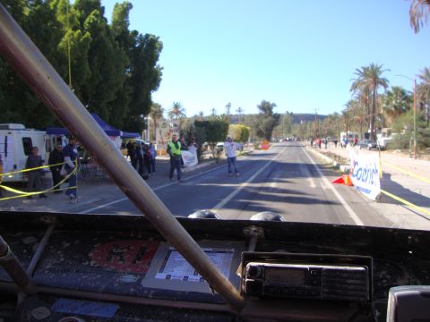 1990 Ford Bronco - view over a dusty dashboard and roll bar shows a palm-lined town street with spectators and banners ahead.