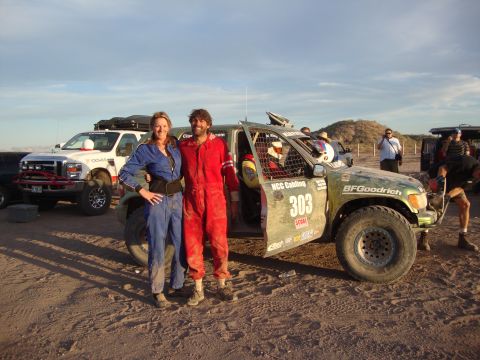 1990 Ford Bronco - couple in racing suits pose beside a mud-covered green race truck numbered 303 with BFGoodrich tires in the desert at dusk.