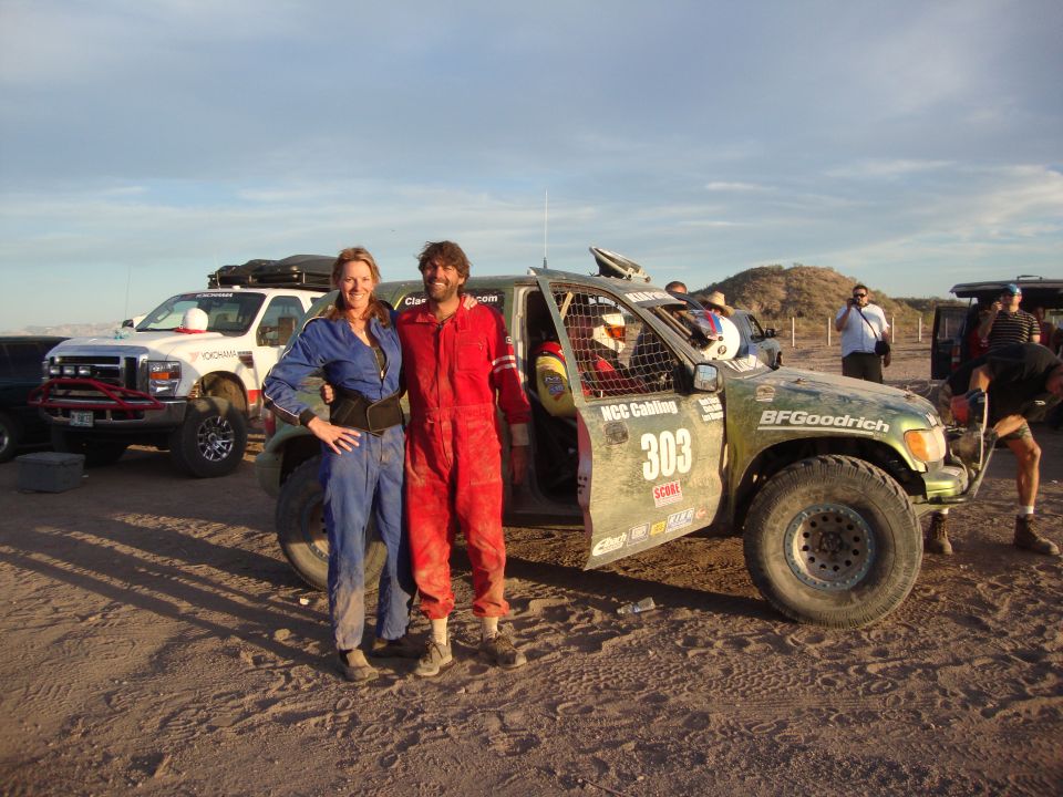 Couple in racing suits pose by mud-covered green race truck 303 in desert at dusk