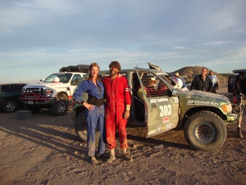 1990 Ford Bronco - couple in blue and red racing suits pose by a green off-road race truck numbered 303, with a white chase truck behind in the desert at dusk.