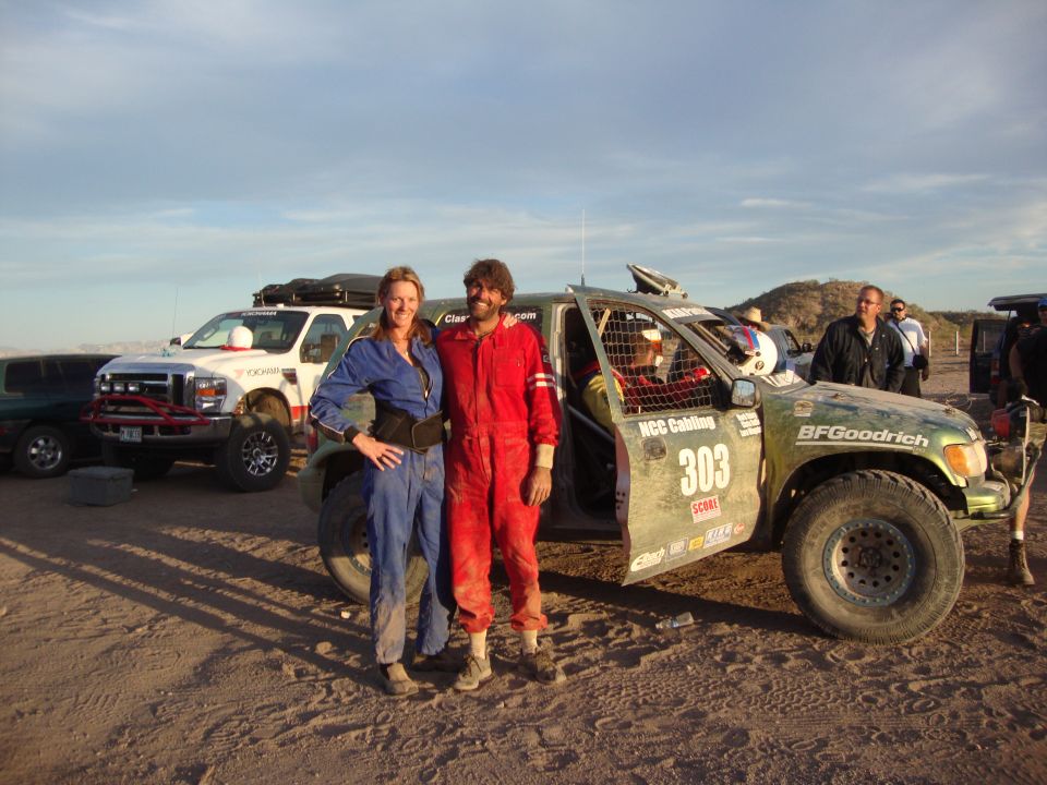 Couple in racing suits pose by green race truck 303 with a chase truck in background