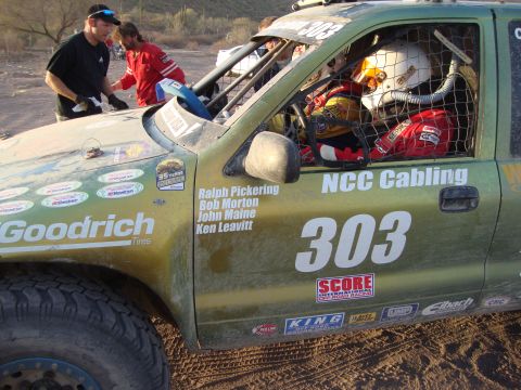 1990 Ford Bronco - Crew members work at the roll-cage opening of a mud-streaked green race truck lettered NCC Cabling and numbered 303 with driver names.