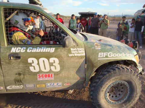 1990 Ford Bronco - helmeted driver sits caged inside a green NCC Cabling race truck numbered 303 as a crowd of spectators looks on in the desert.