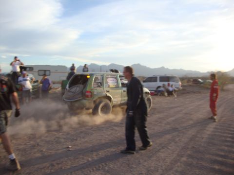 1990 Ford Bronco - man walks past an off-road SUV throwing up dust as spectators and RVs gather at a dusty desert race site near sunset.