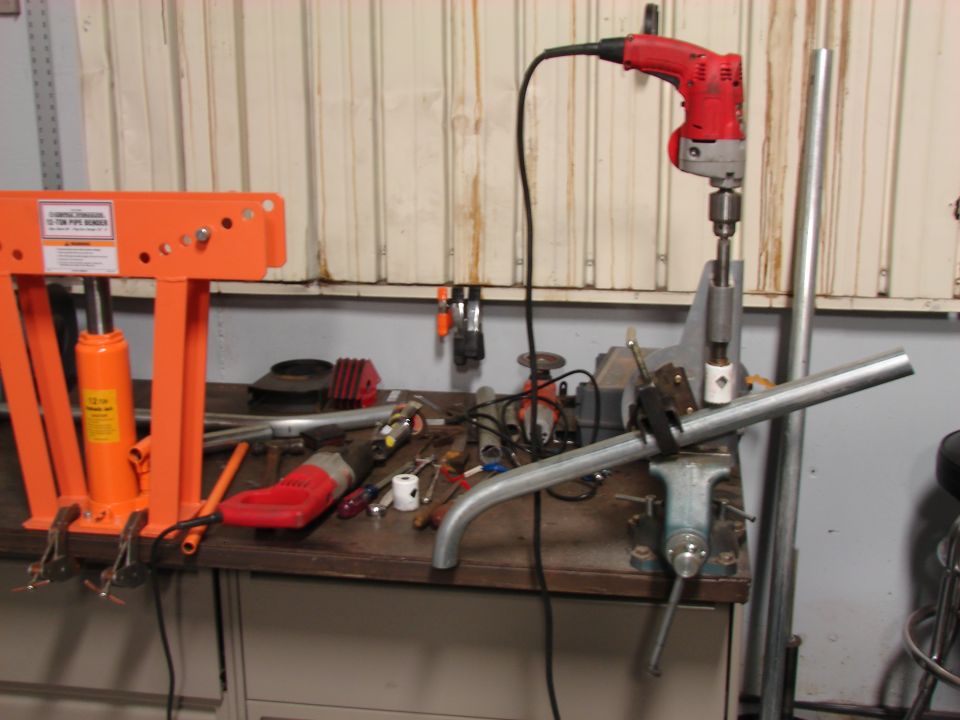 Workbench with pipe bender, drill press, vise, and various tools. A metal pipe is clamped in the vise